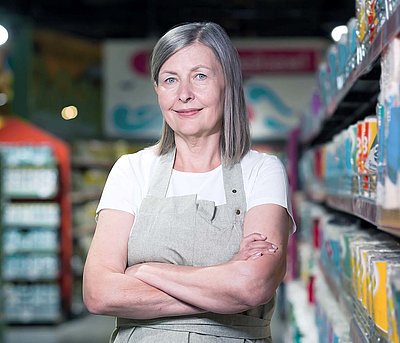 Store managers require a secure and reliable network for their stores Middle-aged store manager with gray long hair and work apron stands between two supermarket shelves smiling contentedly